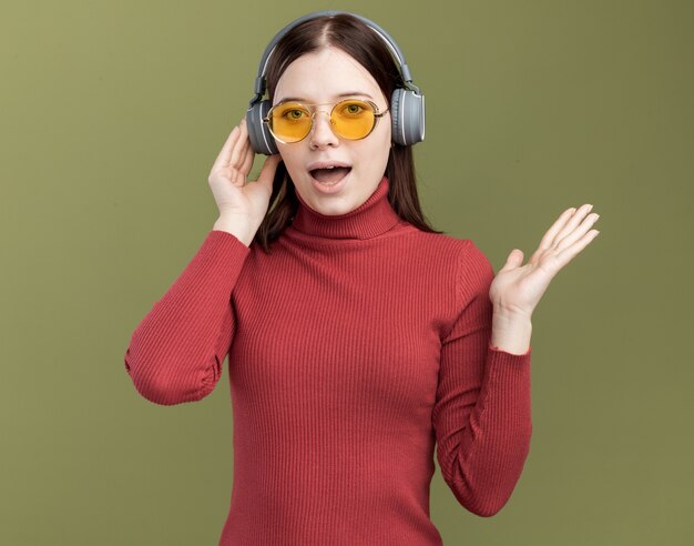 Impresionada joven mujer bonita con gafas de sol y auriculares tocando los auriculares mostrando la mano vacía