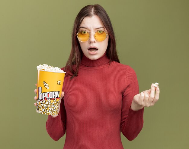 Impresionada joven mujer bonita con gafas de sol sosteniendo un cubo de palomitas de maíz y una pieza de palomitas de maíz mirando al frente aislado en la pared verde oliva