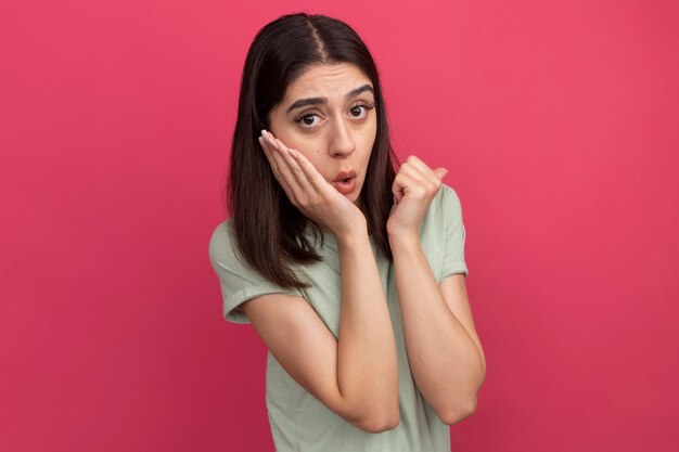 Impresionada joven mujer bonita mirando al frente manteniendo la mano en la cara apuntando hacia atrás aislado en la pared rosa con espacio de copia