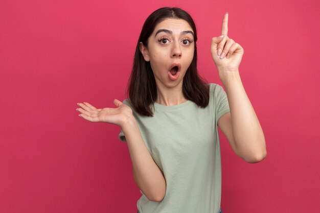 Impresionada joven mujer bonita mirando al frente mostrando la mano vacía apuntando hacia arriba aislado en la pared rosa con espacio de copia