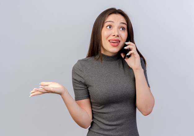 Impresionada joven mujer bonita mirando directamente hablando por teléfono y mostrando la mano vacía aislado sobre fondo blanco.