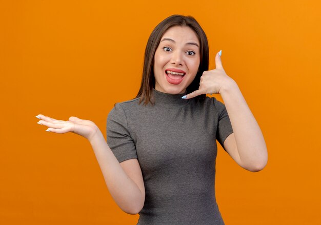 Impresionada joven mujer bonita mostrando la mano vacía y haciendo un gesto de llamada aislado sobre fondo naranja