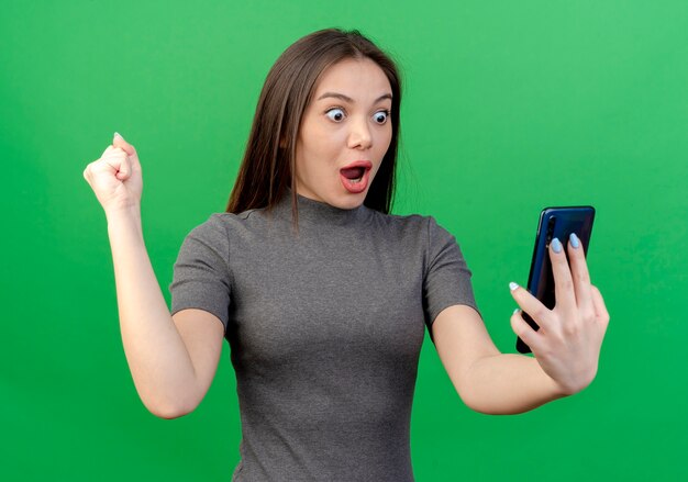 Impresionada joven mujer bonita sosteniendo y mirando el teléfono móvil y apretando el puño aislado sobre fondo verde