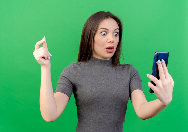 Impresionada joven mujer bonita sosteniendo y mirando el teléfono móvil y manteniendo la mano en el aire aislado sobre fondo verde