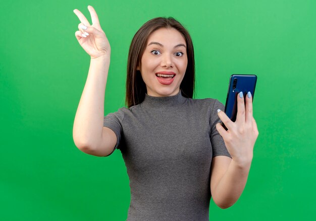 Impresionada joven mujer bonita sosteniendo el móvil y haciendo el signo de la paz teléfono aislado sobre fondo verde