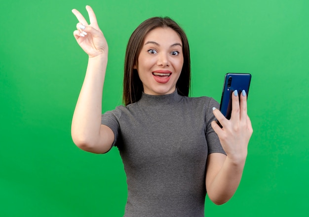 Impresionada joven mujer bonita sosteniendo el móvil y haciendo el signo de la paz teléfono aislado sobre fondo verde