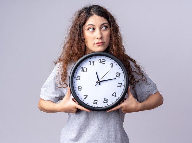 Impresionada joven mujer bonita sosteniendo el reloj mirando al lado aislado en la pared blanca