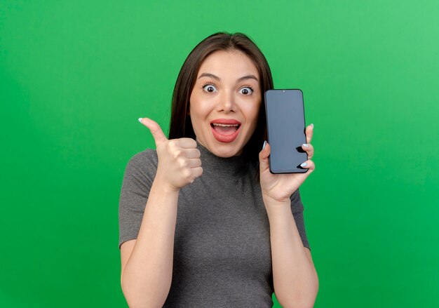 Impresionada joven mujer bonita sosteniendo teléfono móvil y mostrando el pulgar hacia arriba aislado sobre fondo verde con espacio de copia