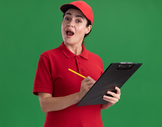 Impresionada joven mujer de entrega en uniforme y gorra sosteniendo portapapeles y lápiz mirando al frente aislado en la pared