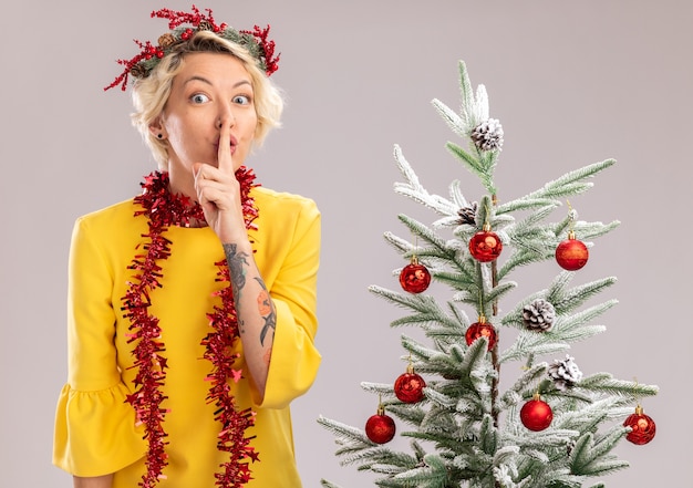 Impresionada joven mujer rubia con corona de Navidad y guirnalda de oropel alrededor del cuello de pie cerca del árbol de Navidad decorado mirando haciendo gesto de silencio aislado en la pared blanca