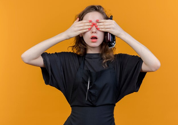 Impresionada joven peluquera eslava vistiendo uniforme sosteniendo cortapelos y cerrando los ojos con las manos aisladas sobre fondo naranja