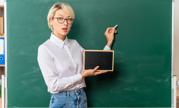 Impresionada joven profesora con gafas en el aula de pie en la vista de perfil delante de la pizarra