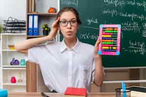Foto gratuita impresionada joven profesora de matemáticas con gafas sentado en el escritorio con útiles escolares mostrando ábaco mirando al frente manteniendo la mano en la cabeza en el aula