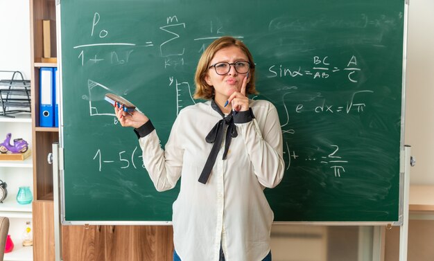 Impresionada joven profesora de pie delante de las herramientas de pizarra sosteniendo el tablero de esponja en el aula