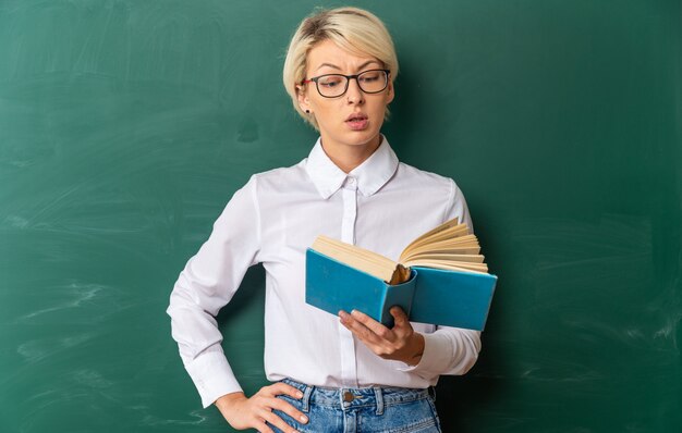 Impresionada joven profesora rubia con gafas en el aula de pie delante de la pizarra sosteniendo y leyendo un libro manteniendo la mano en la cintura