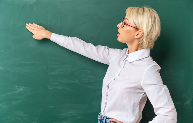 Impresionada joven profesora rubia con gafas en el aula de pie en la vista de perfil frente a la pizarra mirando y apuntando a la pizarra con la mano manteniendo la otra mano en la cintura