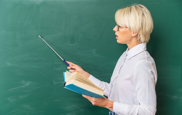 Impresionada joven profesora rubia con gafas en el aula de pie en la vista de perfil frente a la pizarra sosteniendo un libro mirando y apuntando con el puntero en la pizarra