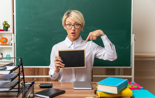 Impresionada joven profesora rubia con gafas sentado en un escritorio con útiles escolares en el aula mostrando y apuntando a la mini pizarra mirando al frente