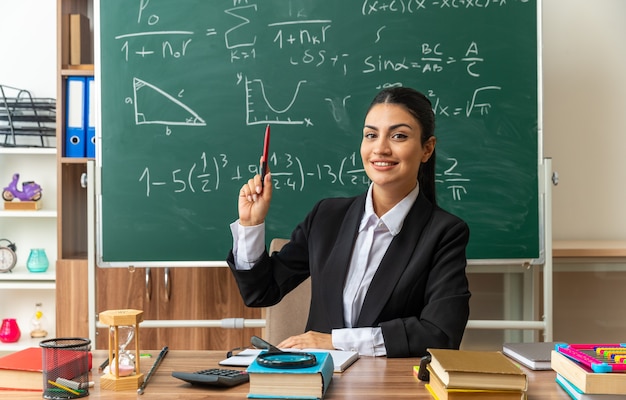 Impresionada joven profesora se sienta a la mesa con herramientas escolares levantando la pluma en el aula