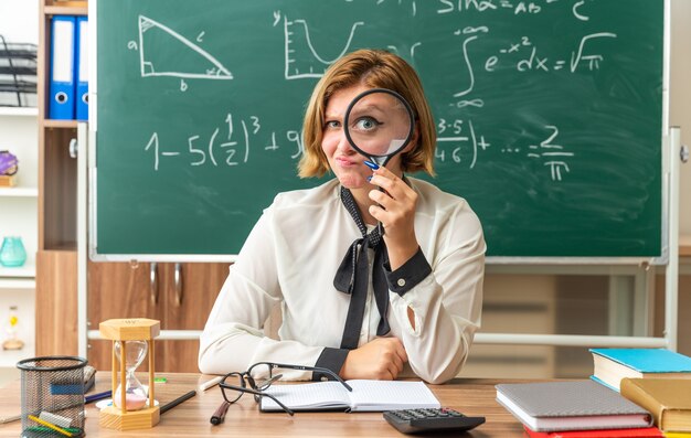 Impresionada joven profesora se sienta a la mesa con herramientas escolares con lupa en el aula