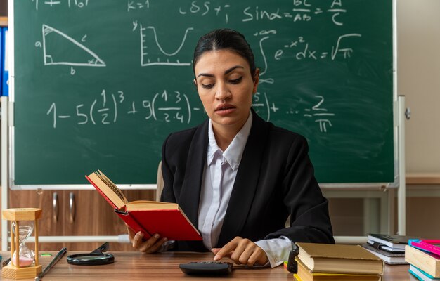 Impresionada joven profesora se sienta a la mesa con útiles escolares sosteniendo el libro y mirando la calculadora en la mano en el aula