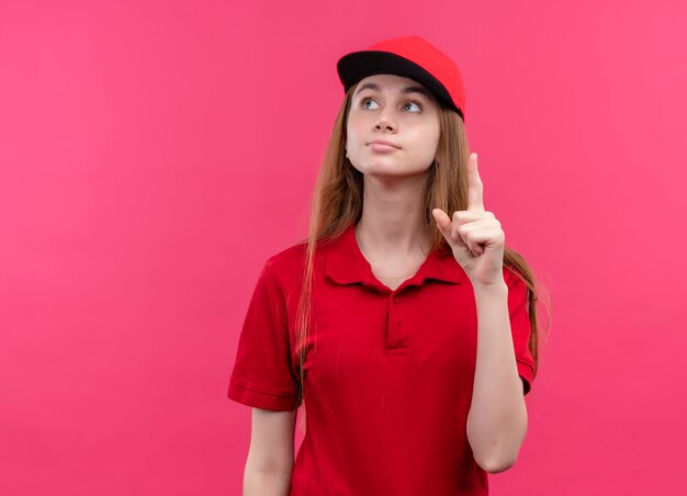 Impresionada joven repartidora en uniforme rojo mirando hacia arriba con el dedo levantado en el espacio rosa aislado con espacio de copia