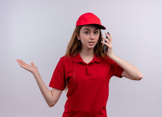 Impresionada joven repartidora en uniforme rojo sosteniendo el teléfono móvil y mostrando la mano vacía en un espacio en blanco aislado