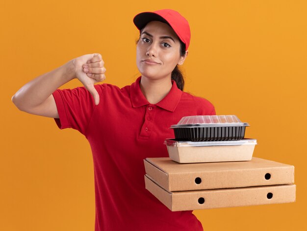 Impresionada joven repartidora vestida con uniforme y gorra sosteniendo cajas de pizza con contenedores de comida mostrando el pulgar hacia abajo aislado en la pared naranja