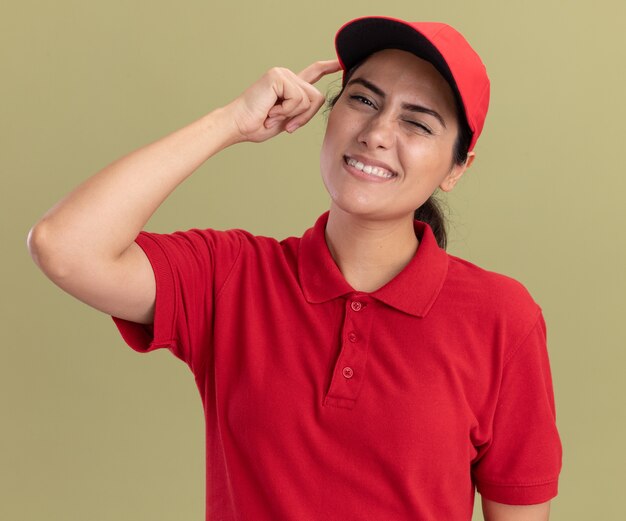 Impresionada joven repartidora vistiendo uniforme con gorra poniendo el dedo en la sien aislado en la pared verde oliva