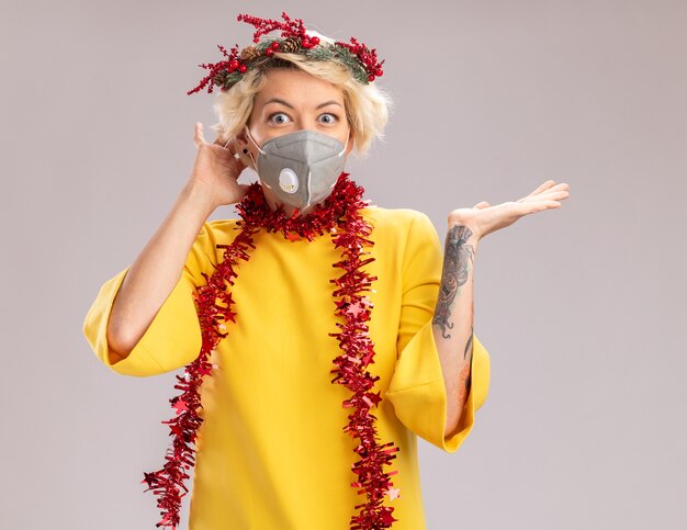 Impresionada joven rubia con corona de Navidad y guirnalda de oropel alrededor del cuello con máscara protectora manteniendo la mano detrás de la cabeza mirando mostrando la mano vacía aislada en la pared blanca