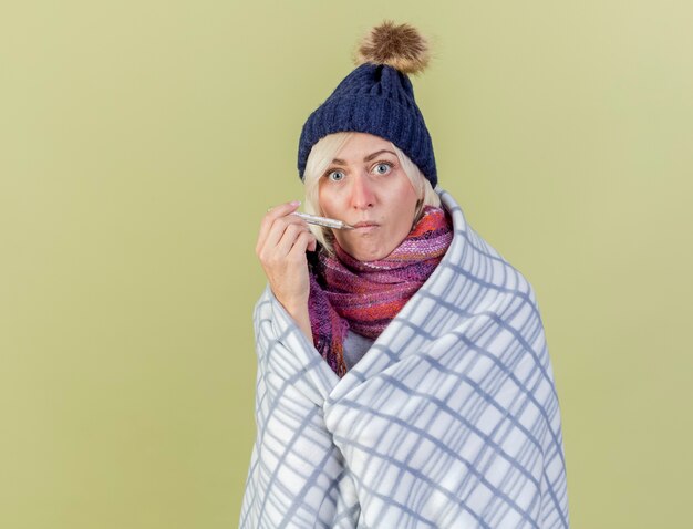 Foto gratuita impresionada joven rubia enferma con gorro de invierno y bufanda envuelta en cuadros pone el termómetro en la boca aislada en la pared verde oliva