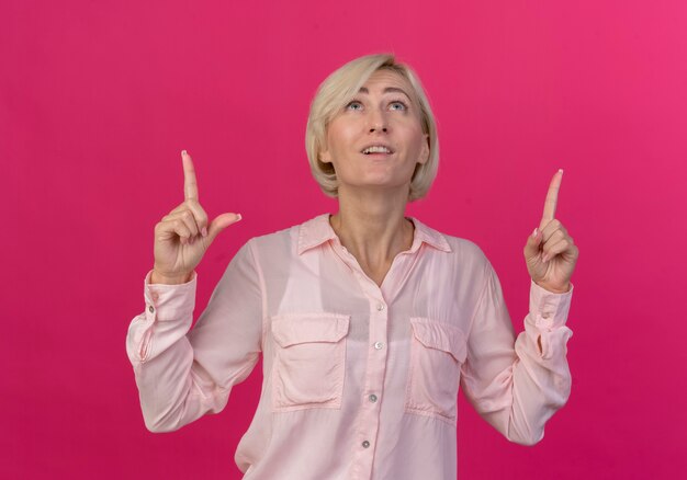 Impresionada joven rubia eslava mirando y apuntando hacia arriba aislado sobre fondo de color rosa