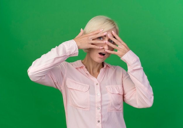 Foto gratuita impresionada joven rubia eslava poniendo los dedos en la cara mirando a la cámara a través de los dedos aislados sobre fondo verde