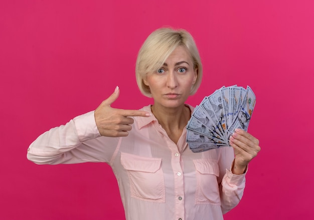 Impresionada joven rubia eslava sosteniendo y apuntando al dinero aislado sobre fondo rosa.