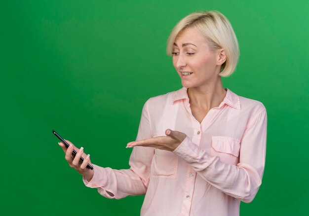 Impresionada joven rubia eslava sosteniendo mirando y apuntando con la mano al teléfono móvil aislado sobre fondo verde