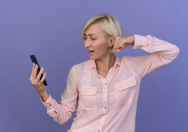 Impresionada joven rubia eslava sosteniendo mirando el teléfono móvil y apretando el puño aislado sobre fondo púrpura
