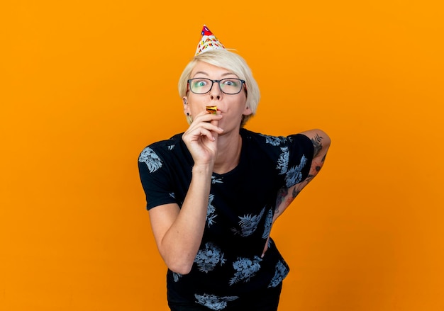 Impresionada joven rubia fiesta con gafas y gorra de cumpleaños manteniendo la mano en la cintura poniendo el soplador de fiesta en la boca mirando al frente aislado en la pared naranja con espacio de copia