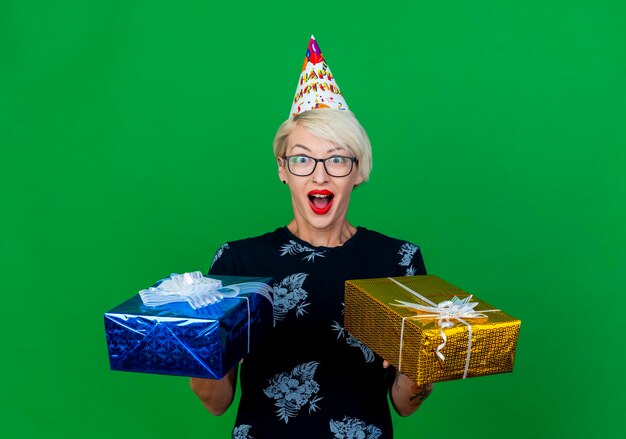 Impresionada joven rubia fiesta mujer con gafas y gorro de cumpleaños sosteniendo cajas de regalo mirando al frente aislado en la pared verde