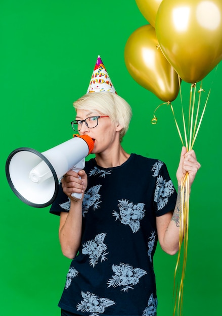 Impresionada joven rubia fiesta mujer con gafas y gorro de cumpleaños sosteniendo globos mirando al frente hablando por altavoz aislado en pared verde