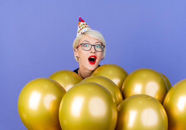 Impresionada joven rubia fiestera con gafas y gorra de cumpleaños de pie detrás de globos mirando a cámara aislada sobre fondo púrpura