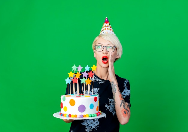 Impresionada joven rubia fiestera con gafas y gorro de cumpleaños sosteniendo pastel de cumpleaños con estrellas mirando a un lado susurrando aislado sobre fondo verde con espacio de copia