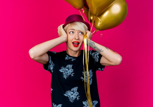 Impresionada joven rubia fiestera con gorro de fiesta sosteniendo globos poniendo las manos en la cabeza mirando al lado aislado sobre fondo carmesí