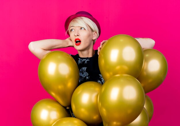 Impresionada joven rubia fiestera con sombrero de fiesta de pie detrás de globos poniendo la mano sobre uno de ellos manteniendo la mano detrás de la cabeza mirando al lado aislado sobre fondo carmesí