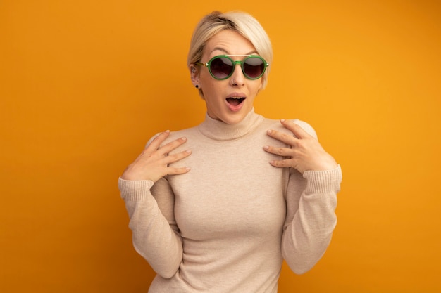 Impresionada joven rubia con gafas de sol poniendo las manos sobre los hombros aislado en la pared naranja con espacio de copia