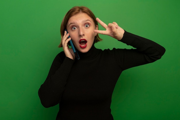 Foto gratuita impresionada joven rubia hablando por teléfono haciendo el signo de la paz aislado en la pared verde