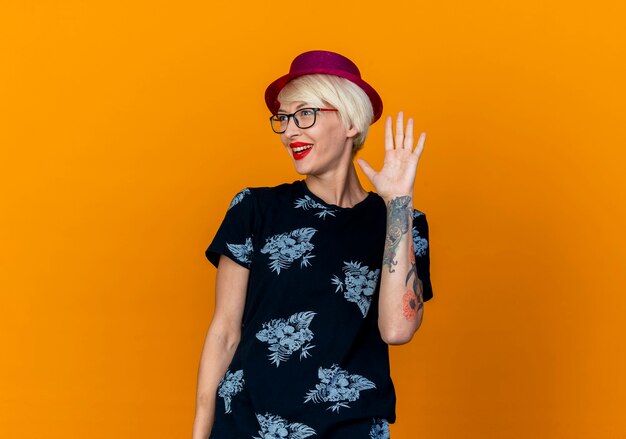 Impresionada joven rubia parte mujer vistiendo gorro de fiesta y gafas mirando al lado haciendo hola gesto aislado en pared naranja