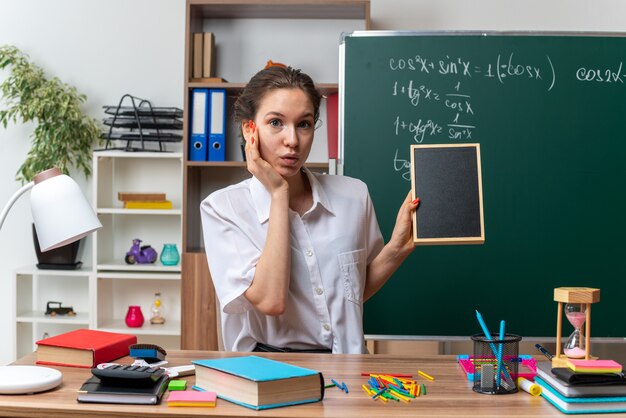 Impresionada joven rubia profesora de matemáticas sentada en un escritorio con herramientas escolares sosteniendo mini pizarra manteniendo la mano en la cara mirando a la cámara en el aula