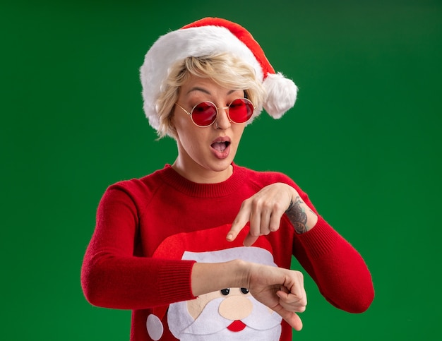 Impresionada joven rubia con sombrero de navidad y suéter de navidad de santa claus con gafas haciendo que eres un gesto tardío aislado en la pared verde