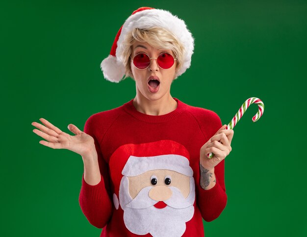 Foto gratuita impresionada joven rubia con sombrero de navidad y suéter de navidad de santa claus con gafas sosteniendo bastón de caramelo de navidad mirando a cámara mostrando la mano vacía aislada sobre fondo verde