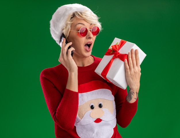 Impresionada joven rubia con sombrero de navidad y suéter de navidad de santa claus con gafas sosteniendo y mirando el paquete de regalo hablando por teléfono aislado en la pared verde